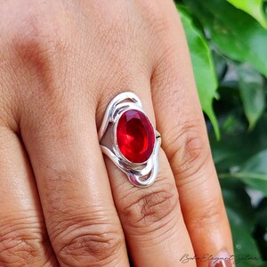 May include: A silver ring featuring a large, oval-cut, deep red gemstone set in a polished metal band. The setting has a swirling, open-work design that frames the central stone, creating a distinct contrast between the vibrant red and the silver.