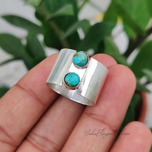 May include: A wide silver band ring featuring two round, bezel-set turquoise stones arranged vertically in the centre. The polished silver surface has a smooth, reflective finish, and the stones display natural blue and brown matrix patterns.