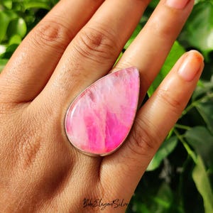 May include: A teardrop-shaped ring featuring a large, polished pink stone with white veining set in a smooth silver bezel. The stone displays a vibrant, translucent pink hue with natural internal patterns and a glossy, reflective surface finish.