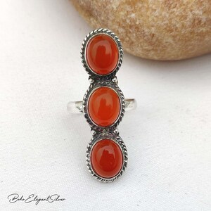 May include: A silver ring featuring three vertically aligned, oval-shaped, smooth orange carnelian stones. Each stone is set within a twisted rope-style silver border. The ring band is plain silver, and the overall design is elongated and symmetrical.