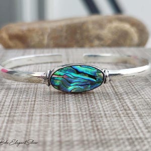 May include: A silver cuff bracelet featuring a central oval-shaped inlay of iridescent abalone shell. The shell displays swirling patterns of deep blue, green, and purple hues. The metal band has a polished finish and an open design at the back.