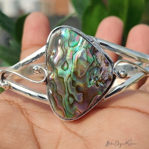 May include: A silver cuff bracelet featuring a large, irregular, polished abalone shell inlay with iridescent green, purple, and blue hues. The metal band displays a smooth, reflective finish with decorative swirling wire accents on both sides.