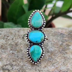 May include: A silver ring featuring three turquoise stones arranged vertically. The top and bottom stones are pear-shaped, while the center stone is oval. Each stone is set in a beaded silver frame, and the band is made of polished silver metal.