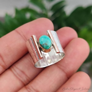 May include: A wide band ring featuring a central oval turquoise stone with brown matrix, set in a gold-toned bezel. The band is made of polished silver with a hammered texture and two vertical gold-toned accents flanking the central stone.