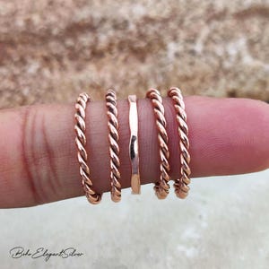 May include: A set of five thin rose gold rings arranged in a row. The set includes four rings with a twisted rope texture and one central ring with a smooth, slightly wavy surface. The metal has a polished, reflective finish throughout the design.