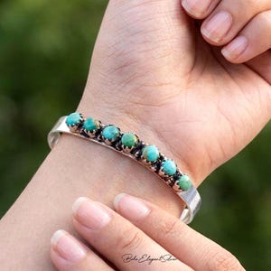 May include: A silver cuff bracelet featuring a row of seven round, polished turquoise stones set in individual scalloped metal bezels. The bracelet has a smooth, polished silver band that wraps around the wrist, showcasing the natural blue-green colour variations of the stones.