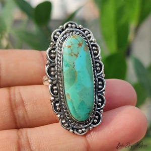 May include: An oval turquoise gemstone ring set in a decorative silver frame. The turquoise stone features natural brown veining and is surrounded by a twisted rope border and a scalloped edge with small silver spheres along the outer perimeter.
