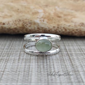 May include: A silver ring featuring a double band design connected by a central oval-shaped, pale green cabochon stone. The polished metal bands create an open space between them, highlighting the smooth, light green gemstone set in the center.