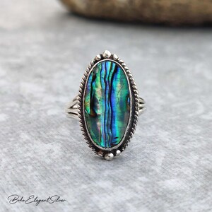May include: An oval-shaped ring featuring a polished abalone shell inlay with iridescent blue and green tones. The shell is set in a silver metal frame with a twisted rope border and small bead accents along the edge of the setting.