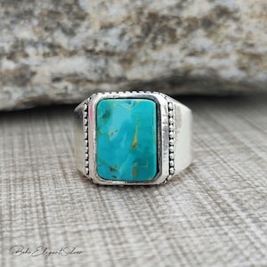 May include: A silver ring featuring a rectangular turquoise stone with brown matrix veins. The stone is set in a smooth bezel, flanked by two vertical rows of small silver beads on either side of the setting, set against a polished silver band.