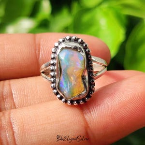 May include: A silver ring featuring an oval-shaped, iridescent opal gemstone with flashes of blue, green, and orange. The stone is set within a beaded metal border, and the band consists of two split silver wires that join at the back of the ring.