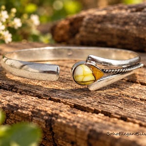 Könnte beinhalten: Ein silberfarbenes Armband mit einer glatten, offenen Schiene und einem tropfenförmigen Stein mit gelber und weißer Maserung. Die Fassung enthält ein gedrehtes Metalldetail in der Nähe des Steins. Der Text BohoElegantSilver ist sichtbar.