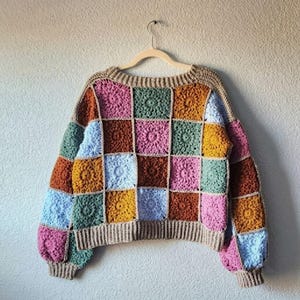 May include: A long-sleeve sweater constructed from multi-colored crochet granny squares in shades of pink, green, mustard yellow, brown, and light blue. The garment features a tan ribbed neckline, cuffs, and hem, and is displayed on a hanger.