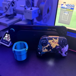 May include: A black handheld gaming console, a wireless game controller featuring a yellow Pikachu and purple Mewtwo graphic, and a small, cylindrical, metallic blue plastic cup-shaped accessory sitting on a dark textured surface.