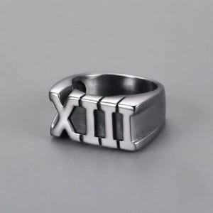 Op de afbeelding: Een gepolijste donkerzilverkleurige metalen ring met het Romeinse cijfer XIII in reliëf op de voorzijde. De band heeft een glad, afgerond profiel met een metallic afwerking, waarbij de verhoogde tekens duidelijk afsteken tegen de achtergrond.