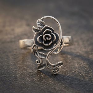 Peut inclure: Une bague argentée ornée d'un motif de rose proéminent avec des pétales et des feuilles détaillés. Le design comprend une tige incurvée qui s'étend verticalement depuis l'anneau, présentant une finition oxydée soulignant les textures florales.