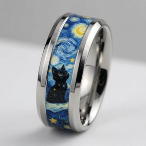 Op de afbeelding: Een gepolijste zilverkleurige metalen ring met een centrale inleg met een blauw en geel wervelend nachthemelpatroon. Een kleine zwarte kat met grote ogen is afgebeeld zittend in de sterrenhemel, omringd door gele sterren en wolken.