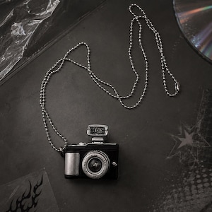 Peut inclure: Un pendentif miniature en forme d'appareil photo noir et argenté, suspendu à une longue chaîne à billes en argent. L'appareil présente un objectif avec l'inscription 1:4.8 55mm OPTICALS et un petit flash rectangulaire sur le dessus.