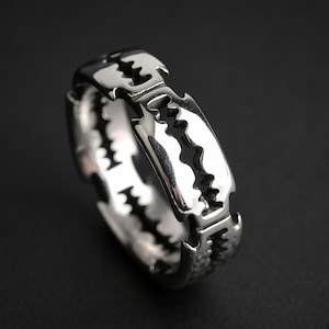 Peut inclure: Une bague en métal argenté poli présentant un design en anneau continu qui imite la forme d'une lame de rasoir. La bague possède une surface réfléchissante avec des détails découpés sur les bords, créant un aspect dentelé et irrégulier.