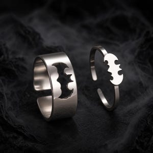Puede incluir: Dos anillos de metal plateado con un diseño recortado en forma de murciélago. El anillo más grande tiene una banda ancha con una silueta de murciélago en negativo, mientras que el más pequeño tiene una banda fina con un centro de murciélago.