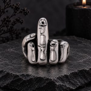 Op de afbeelding: Een zilverkleurige metalen ring in de vorm van een hand die een middelvingergebaar maakt. Het ontwerp bevat gedetailleerde knokkels, vingernagels en huidplooien met een donkere, geoxideerde afwerking in de groeven om de vingeranatomie te benadrukken.