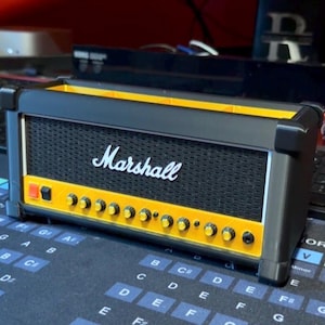 May include: A black and yellow Marshall amplifier-style desk organizer featuring a black mesh front panel with the white Marshall logo, a yellow control panel with small knobs, and a rectangular open top compartment for storing office supplies.