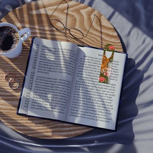 Puede incluir: Un libro abierto con un marcapáginas que presenta una ilustración de un tigre y motivos florales rojos, colocado sobre una mesa de madera junto a una taza blanca con café negro y unas gafas de montura fina negra sobre las páginas.