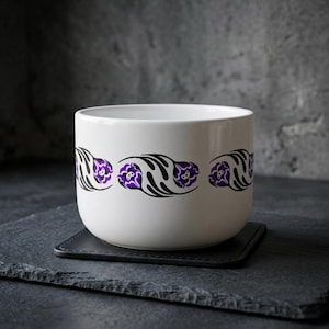Puede incluir: Un cuenco de cerámica blanca con una banda horizontal repetitiva de motivos florales estilizados en tonos morado y negro. El cuenco tiene un exterior liso y redondeado con una abertura amplia, mostrando un diseño limpio y uniforme.