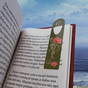 Puede incluir: Un marcapáginas de color verde oliva con la parte superior redondeada y un orificio, que presenta un diseño floral vertical con flores rojas y contornos de enredaderas en blanco. El marcapáginas está colocado dentro de un libro abierto.