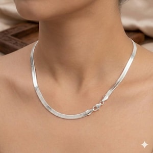 Puede incluir: Un collar de cadena de espiga en tono plateado con un diseño de eslabones planos y flexibles que descansa sobre el cuello. El collar incluye un mecanismo de cierre de langosta seguro visible en el lateral, con un acabado metálico pulido.