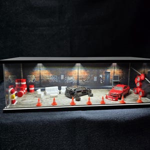 Może przedstawiać: Miniaturowa gablota dioramowa garażu z czarnym samochodem sportowym, czerwonym samochodem sportowym, białymi meblami wypoczynkowymi, czerwono-białymi beczkami i pomarańczowymi pachołkami drogowymi na tle ściany z cegły z oświetleniem.