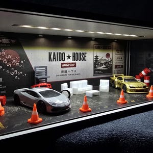 May include: A miniature diorama display case featuring a silver sports car and a gold sports car parked on a dark surface. The scene includes orange traffic cones, white miniature furniture, and a backdrop with Kaido House branding and Japanese text.