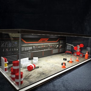 Może przedstawiać: Miniaturowa diorama garażu Formuły 1 w przezroczystej akrylowej gablocie. Wnętrze zawiera grafikę ścienną z narzędziami, czerwono-białe beczki, pomarańczowe pachołki drogowe, czarne opony, białe meble oraz czerwony stojak na koła.