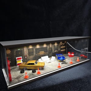 Może przedstawiać: Miniaturowa witryna diorama garażu z dwoma samochodzikami, złotym i niebieskim, białymi meblami, pomarańczowymi pachołkami drogowymi i czarnymi oponami. Tylna ściana przedstawia nadrukowane narzędzia warsztatowe i znaki, oświetlone lampkami.