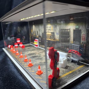 Może przedstawiać: Miniaturowa diorama garażu zamknięta w przezroczystej akrylowej obudowie. Wnętrze zawiera nadrukowaną ścianę z tekstem "WORK HARD STAY HUMBLE", "EXIT" oraz "FIRE EXIT". Na podłodze znajdują się żółte pasy ostrzegawcze, pomarańczowe pachołki drogowe, białe meble i czerwony zderzak samochodowy.