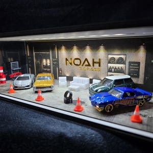 May include: A miniature garage diorama titled NOAH GARAGE featuring a concrete floor, LED lighting, and several die-cast toy cars including a silver sports car, a gold sedan, a dark green hatchback, and a blue muscle car with orange traffic cones.