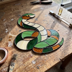 May include: A stained glass snake ornament featuring a coiled body constructed from individual pieces of green, amber, and cream-coloured glass held together by dark metal solder lines, with a distinct head shape and a single dark eye detail.