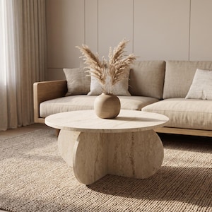 May include: A round beige stone coffee table with a thick, curved cross-shaped base. A small, smooth, spherical beige vase sits on the tabletop, containing a cluster of dried, feathery beige pampas grass stems.