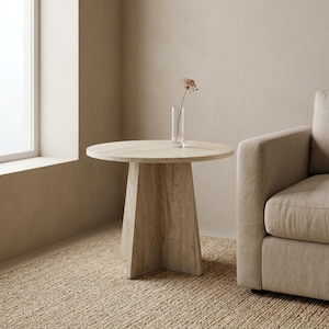 Puede incluir: Una mesa auxiliar redonda con acabado en piedra travertino de color beige claro y una base central de pedestal. Sobre la superficie de la mesa, que presenta vetas naturales, hay un jarrón cilíndrico de cristal con una flor seca.