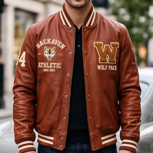 Può includere: Una giacca varsity in pelle marrone con polsini e colletto a coste a righe color crema. La parte anteriore presenta la scritta BACKAVEN ATHLETIC SINCE 1984 con un grafico di un lupo, il numero 84 sulla manica e una grande lettera W con WOLF PACK.
