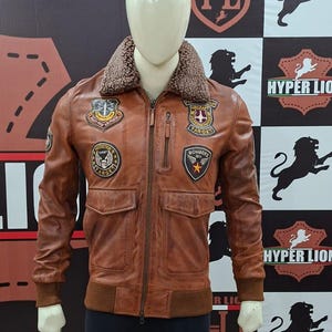 Puede incluir: Una chaqueta bomber de cuero marrón con cuello de piel sintética marrón, cierre de cremallera frontal y puños acanalados. La chaqueta está decorada con varios parches bordados en el pecho, incluyendo el texto "EIGHTY FOUR AIR FORCE" y "BOMBERS DIV".