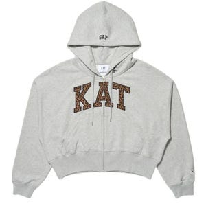 Puede incluir: Una sudadera con capucha y cremallera en color gris claro que presenta la palabra KAT en letras con estampado de leopardo marrón y negro en el pecho. La prenda incluye una capucha con la palabra GAP bordada en negro y un dobladillo corto.