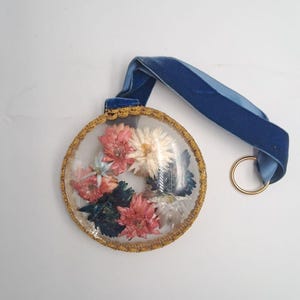 Pode incluir: Um ornamento circular com uma cúpula transparente preenchida com flores secas cor-de-rosa, brancas e azul-escuras. A borda é rematada com fio metálico dourado. Uma fita de veludo azul está presa ao topo, terminando num pequeno anel metálico dourado.
