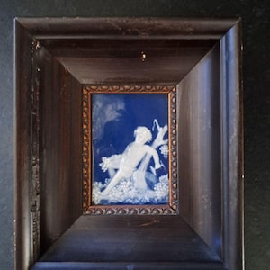 Può includere: Una cornice rettangolare in legno marrone scuro racchiude una piccola placca blu profondo con un rilievo bianco di una figura distesa circondata da fogliame. Il bordo interno della cornice presenta un motivo testurizzato color rame.