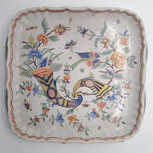 Peut inclure: Un plat carré en céramique aux bords festonnés, orné d'un motif peint à la main représentant deux cornes d'abondance, des fleurs colorées, du feuillage vert et un oiseau. La bordure présente un motif quadrillé rouge sur fond blanc.
