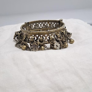Peut inclure: Un bracelet jonc métallique doté d'une large bande avec un motif en treillis découpé et une bordure en corde torsadée. Le bord inférieur est orné d'une série de breloques pendantes, incluant de petites clochettes sphériques et des pendentifs plats.