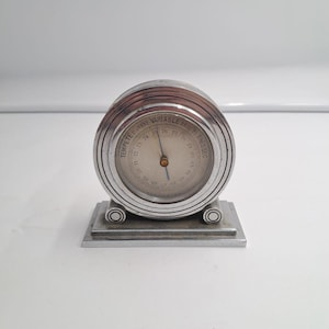 Puede incluir: Un barómetro circular de metal plateado con una base rectangular escalonada. La esfera presenta una aguja central y texto impreso que incluye "TEMPETE", "VARIABLE" y "TRES SEC", junto con marcas numéricas que van del 69 al 83.