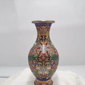 May include: A metal vase featuring intricate cloisonné enamel work with a gold-toned base. The surface is decorated with colorful floral and scrolling patterns in shades of blue, green, red, and white, finished with a smooth, rounded metal rim.