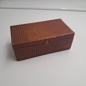 Könnte beinhalten: Eine rechteckige Aufbewahrungsbox mit brauner Außenseite und einer strukturierten Krokodilleder-Optik. Die Box verfügt über einen Klappdeckel und einen kleinen, mittig platzierten Metallverschluss an der Vorderkante. Das Design ist schlicht.