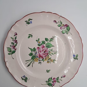Peut inclure: Une assiette en céramique ronde avec un bord festonné et un liseré rouge peint. Le centre présente un bouquet floral peint à la main avec une grande rose rose, des fleurs jaunes et des feuilles vertes. Des motifs floraux ornent le bord.